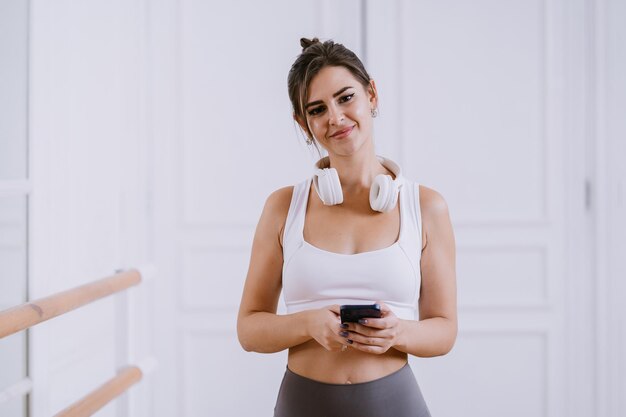 Atractiva chica estadounidense morena en ropa deportiva y auriculares en el cuello sostiene el teléfono mira a la cámara