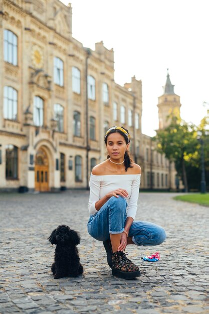 Atractiva chica hispana sentada en la calle con un caniche de juguete de raza de perro y posando para la cámara