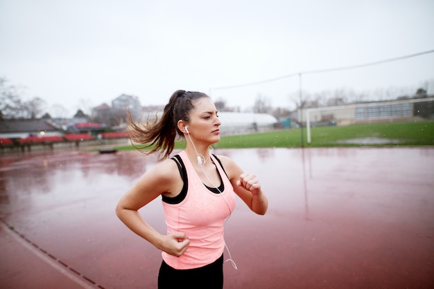 Atractiva chica joven y motivada que se apresura decisivamente hacia el final de su carrera cerca del campo de fútbol mientras llueve.