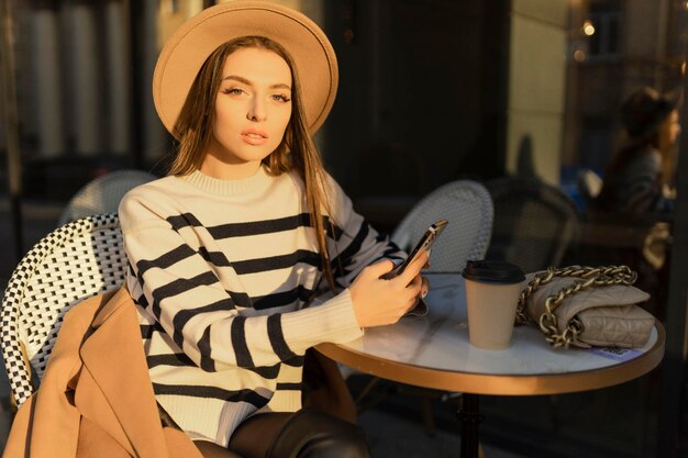 Atractiva chica morena alegre en gabardina con café para ir felizmente usando el teléfono celular al aire libre