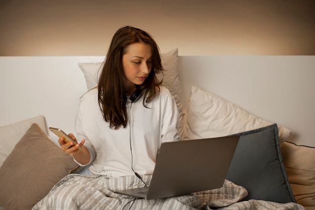 Atractiva chica morena sentada en la cama con su teléfono y computadora portátil en un día vago