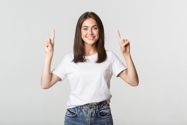 Atractiva chica morena sonriente apuntando con el dedo hacia arriba, mostrando el logo.