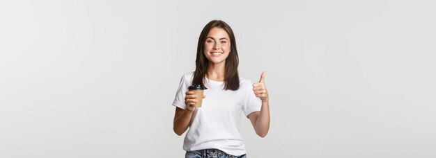 Atractiva chica morena sonriente que parece satisfecha bebiendo café y muestra el pulgar hacia arriba recomendar café