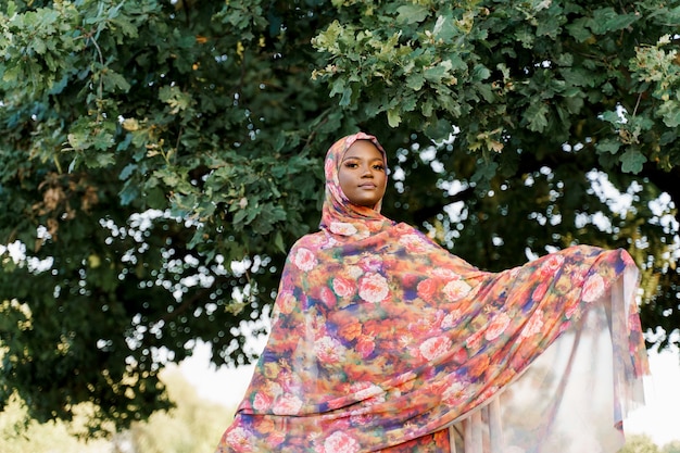 Atractiva chica negra musulmana vestida con hiyab sonríe cerca de árbol verde