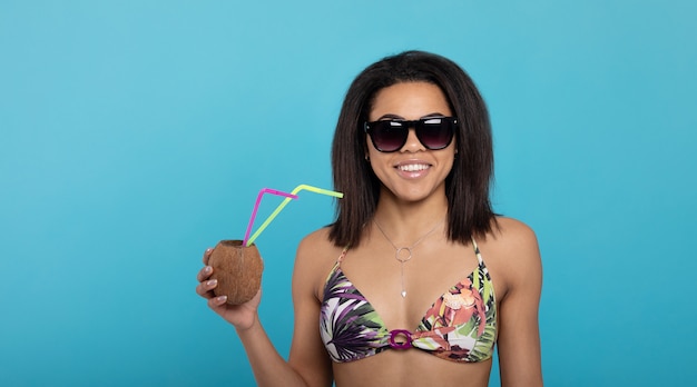 Atractiva chica negra en traje de baño y gafas de sol con cóctel de verano y sonriendo a la cámara.