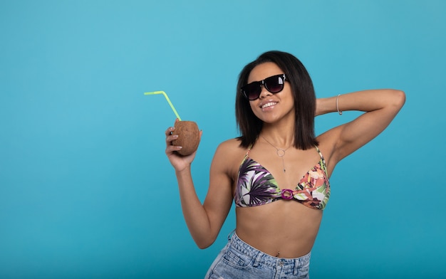 Atractiva chica negra en traje de baño y gafas de sol con cóctel de verano y sonriendo a la cámara.