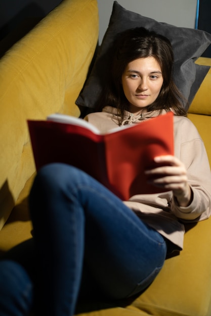 Foto atractiva chica de pelo largo lee atentamente el libro acostado en un cómodo sofá con las rodillas dobladas