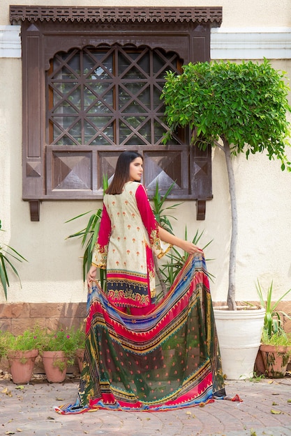 Atractiva chica en pose trasera con vestido Desi sosteniendo Dupatta para disparar al aire libre con plantas