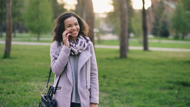 Atractiva chica de raza mixta hablando de teléfono inteligente y bebiendo café en el parque de la ciudad con bolsas