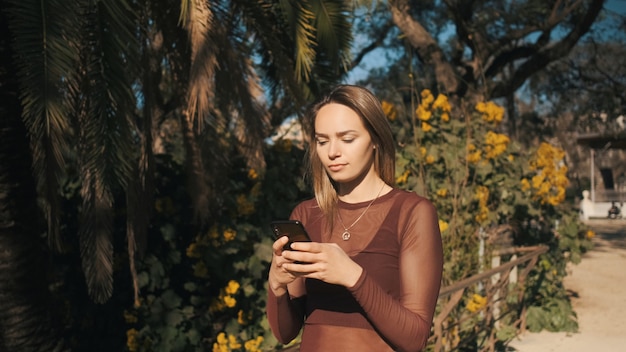 Atractiva chica rubia con atención usando smartphone en el parque de la ciudad