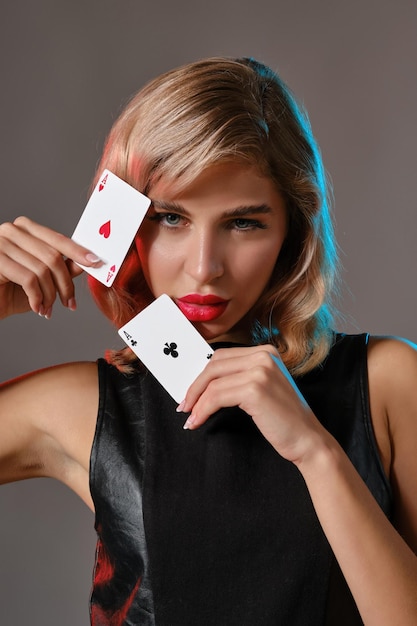 Atractiva chica rubia con maquillaje brillante en vestido de cuero negro está mostrando dos naipes y posando contra el fondo gris del estudio Concepto de casino de póquer de entretenimiento de juego Primer plano