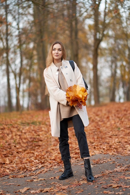 Atractiva chica rubia con ramo de hojas de arce naranja en las manos con abrigo de otoño de moda, jeans negros y botas de cuero negro de moda caminando en el parque de otoño.