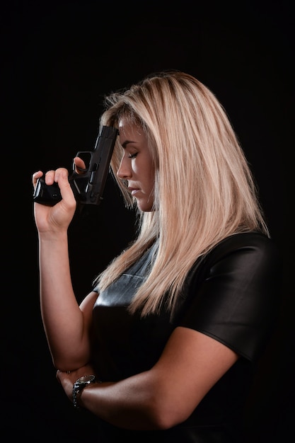 Atractiva chica rubia sosteniendo pensativamente una pistola negra sobre un fondo muy oscuro en ropa negra. Estudio de rodaje