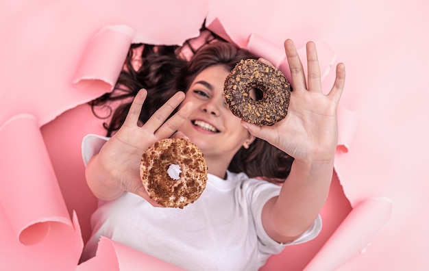 Atractiva chica sonriente con apetitosas donas mira desde un agujero en un fondo de papel rosa, vista superior.