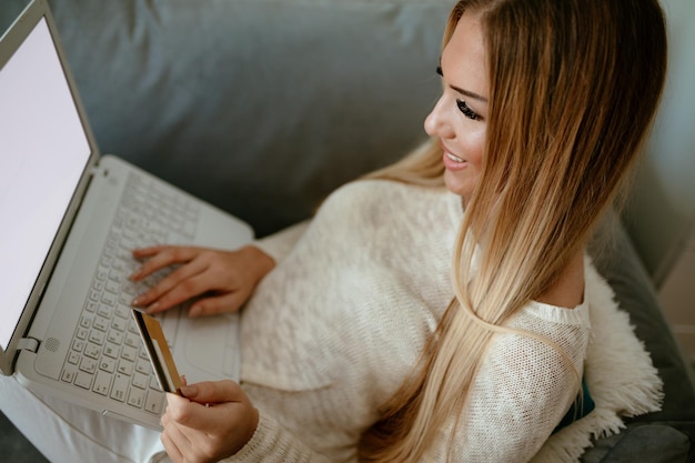 Atractiva chica sonriente sentada en su sala de estar con tarjeta de crédito pagando cuentas por computadora portátil. Enfoque selectivo. Vista superior.