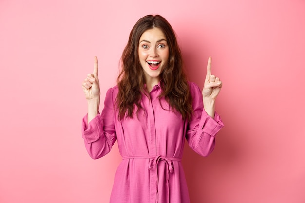 Atractiva dama feliz apuntando con el dedo hacia arriba y sonriendo alegre, mostrando un anuncio impresionante, de pie sobre una pared rosa.