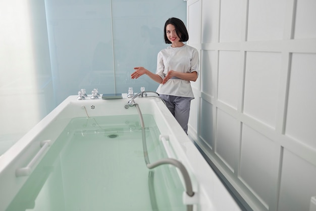 Atractiva dama en uniforme invita a tomar un baño de hidromasaje contemporáneo lleno de agua clara