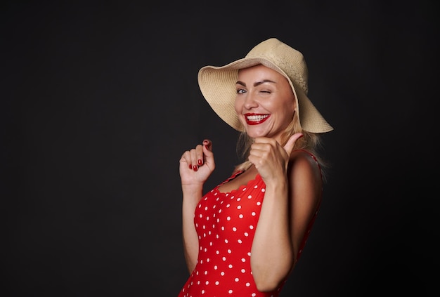 Atractiva y deslumbrante mujer rubia con sombrero de paja de verano y traje de baño rojo guiñando un ojo y sonriendo con una hermosa sonrisa blanca mostrando el pulgar hacia la cámara posando sobre fondo negro con espacio para copiar