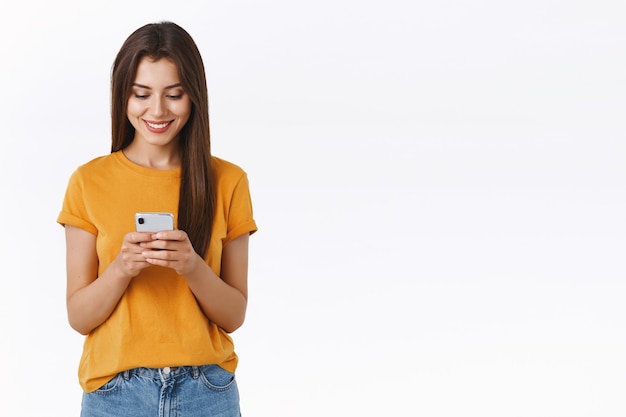 Atractiva, despreocupada joven mujer morena emotiva en camiseta amarilla, sosteniendo un teléfono inteligente sonriendo emocionado, mirando la pantalla del móvil, haciendo el pedido, haciendo compras el viernes negro, soporte fondo blanco