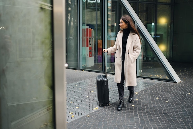 Atractiva y elegante mujer de negocios con abrigo esperando en la calle con una pequeña maleta