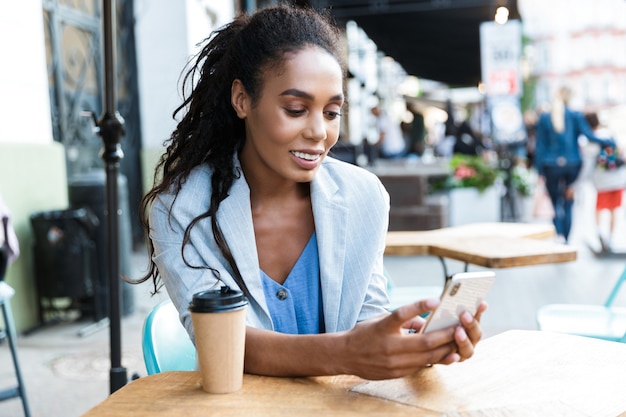 Atractiva empresaria africana joven sonriente sentada en la mesa de café al aire libre, utilizando el teléfono móvil mientras bebe café en una taza para llevar