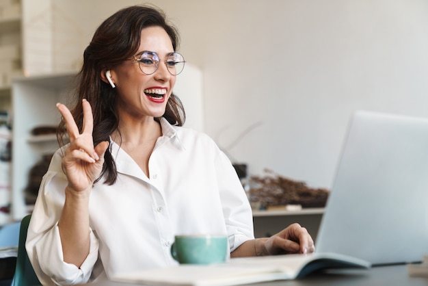 Atractiva empresaria morena joven alegre sentada en la mesa de café con computadora portátil en el interior, usando auriculares inalámbricos, llamada de audio, gesto de paz