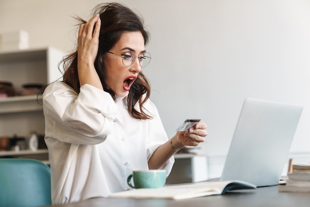 Atractiva empresaria morena joven molesta sorprendida sentada en la mesa de café con computadora portátil en el interior, compras en línea con tarjeta de crédito