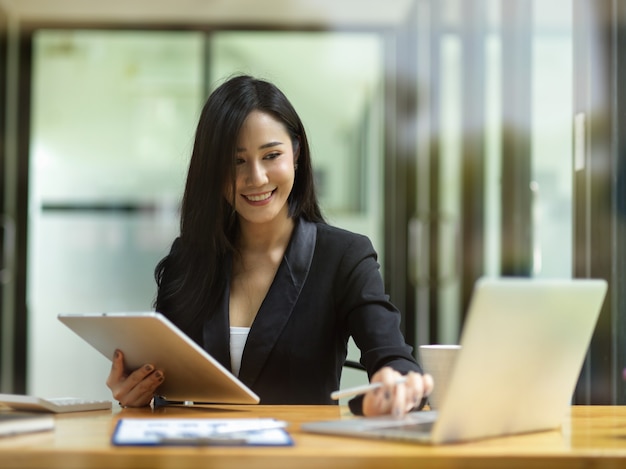 Atractiva empresaria en traje negro usando una computadora portátil y sosteniendo una tableta inteligente para verificar datos