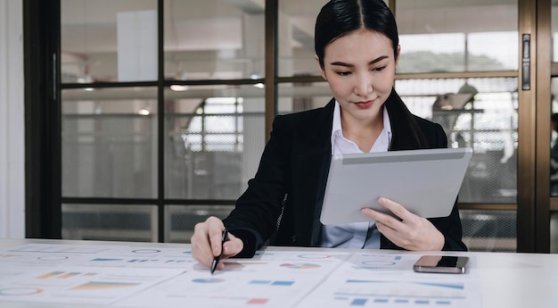 Atractiva y encantadora joven empresaria asiática o trabajadora financiera con traje formal usando tableta y trabajando con su informe financiero en la oficina