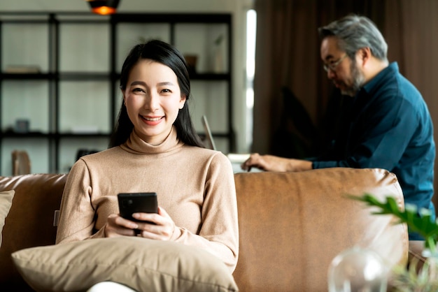 Atractiva esposa asiática sostiene el teléfono inteligente en el sofá en la sala de estar con el esposo trabajando desde casa con una computadora portátil sentada en la mesa de comedor detrás del concepto de estilo de vida del trabajo desde el hogar