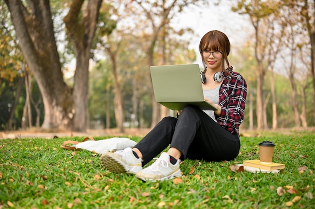 Atractiva estudiante universitaria asiática usando su computadora portátil mientras se sienta en el césped en el parque