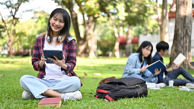 Atractiva estudiante universitaria asiática usando su tableta mientras se relaja sentada en el parque del campus