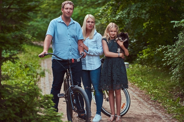 Una atractiva familia vestida con ropa informal en un paseo en bicicleta con su lindo perrito spitz, de pie en el parque.