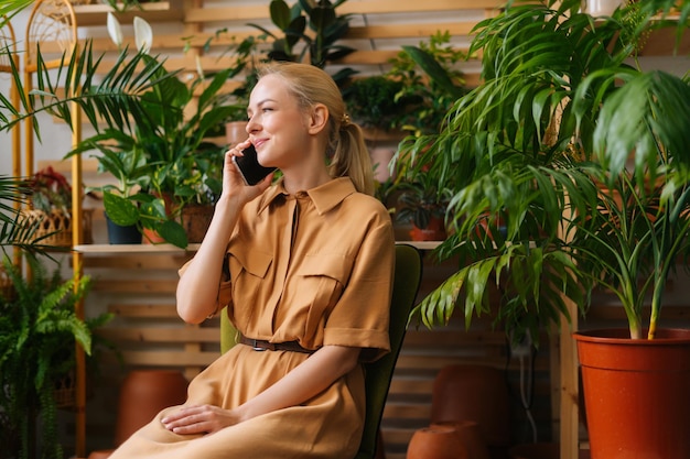 Atractiva y feliz dueña de una tienda de flores sentada en su propia tienda de flores y hablando por teléfono inteligente con el cliente