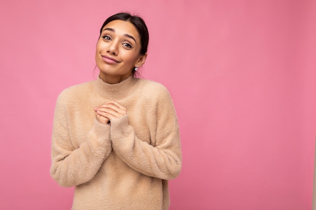 Atractiva feliz linda adorable tierna joven morena mujer en suéter beige casual aislado sobre fondo rosa con espacio libre y disfrutando