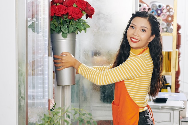 Atractiva floristería asiática sonriente tomando un balde de rosas de adorno floral