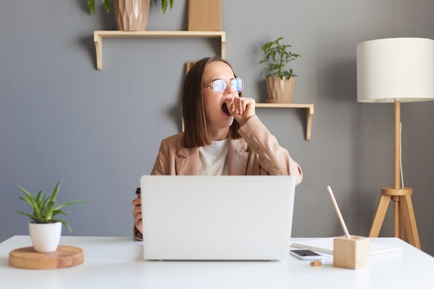 Atractiva y hermosa mujer caucásica joven que se siente cansada y sobrecargada de trabajo con anteojos trabaja de forma remota desde casa o sentada en la mesa en la oficina frente a la computadora portátil bostezando y cubriendo la boca