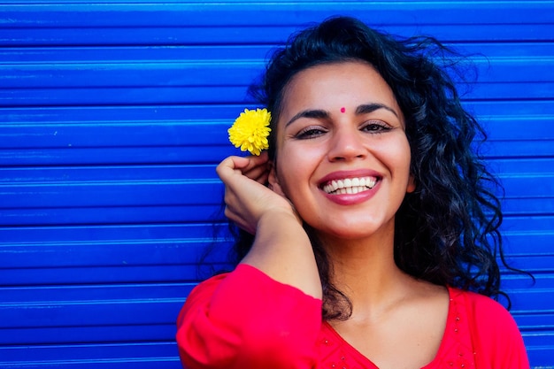 Atractiva hermosa mujer hispana latina feliz joven con bindi de teca en la frente sonriendo sobre fondo azul de Wall Street sosteniendo flover fresco