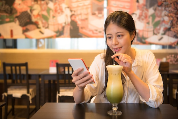 atractiva y hermosa turista sentada en la famosa tienda de bebidas de Japón y usando el sitio web de la comunidad de navegación de teléfonos celulares cuando prueba la leche de té verde matcha helado japonés.