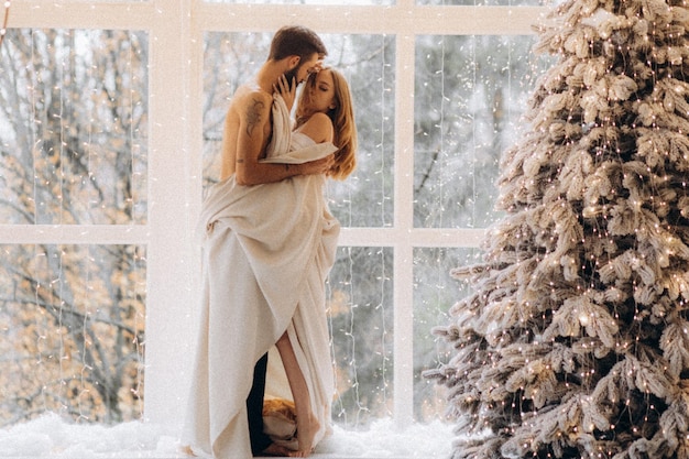 atractiva joven abrazando a un novio sin camisa cerca del árbol de navidad en casa