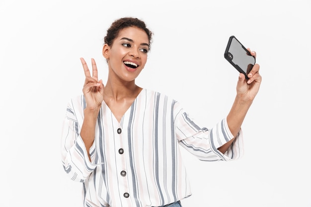 Atractiva joven africana con ropa casual que se encuentran aisladas sobre una pared blanca, tomando un selfie, mostrando la paz