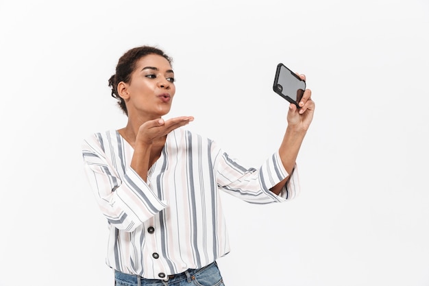 Atractiva joven africana vistiendo ropa casual que se encuentran aisladas sobre una pared blanca, tomando un selfie, enviando un beso