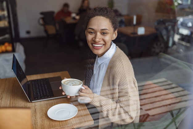 Atractiva joven afroamericana sosteniendo una taza con capuchino disfrutando del trabajo remoto en una laptop en una cafetería