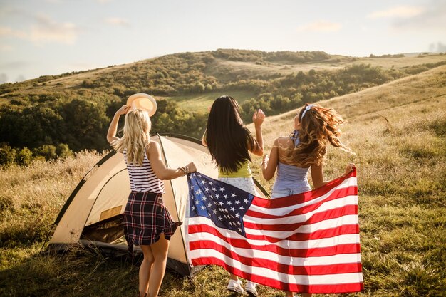 Una atractiva joven amiga con bandera estadounidense disfrutando y festejando frente a una carpa en el campamento.