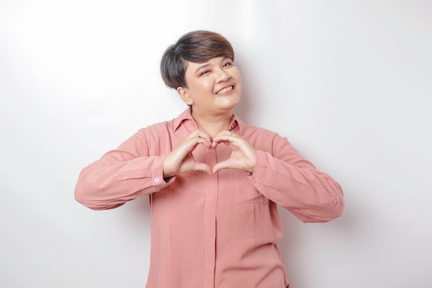 Una atractiva joven asiática de pelo corto con una camisa rosa se siente feliz y un gesto romántico del corazón expresa sentimientos tiernos