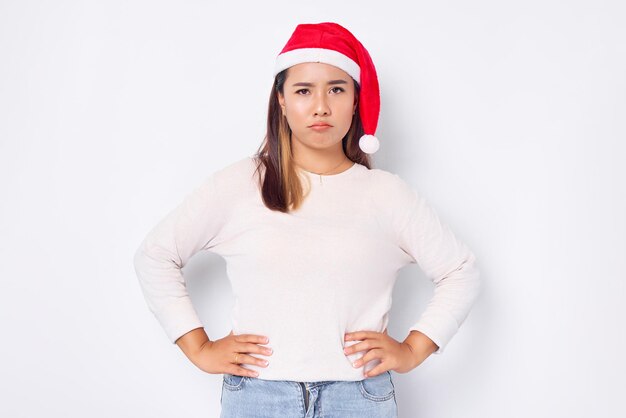 Atractiva joven asiática con sombrero de Papá Noel tomándose de la mano en la cintura y expresión de enojo ante la cámara aislada sobre el fondo blanco