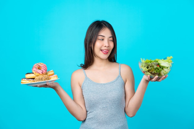 Atractiva joven asiática sosteniendo y eligiendo entre disco de donas o ensalada de verduras en un tazón de vasos