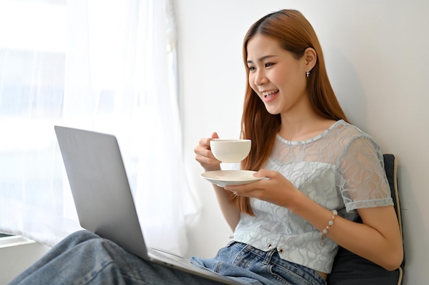 Atractiva joven asiática trabajando remotamente en el café tomando un café usando una laptop
