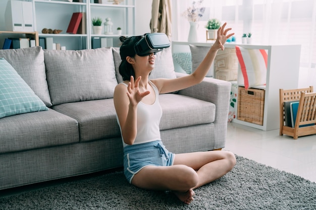 Atractiva joven con auriculares VR gesticulando y sonriendo mientras se sienta en el salón de casa en el suelo. hermosa mujer china asiática se relaja en la alfombra con gafas de realidad virtual jugando.