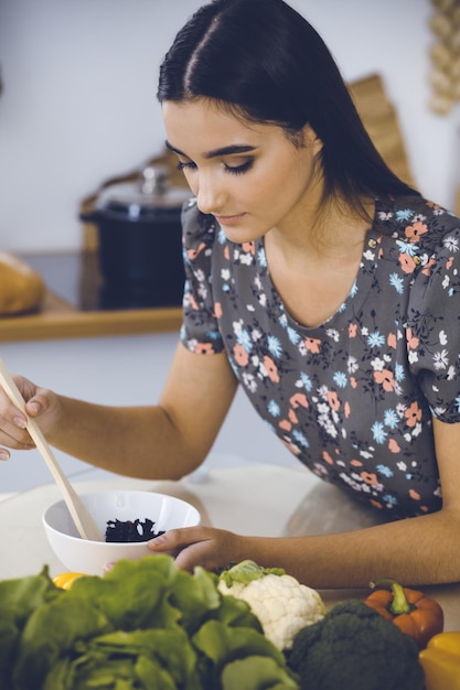 Foto una atractiva joven de cabello oscuro prueba una nueva receta para una deliciosa mezcla de ensaladas mientras se sienta en la mesa de la cocina